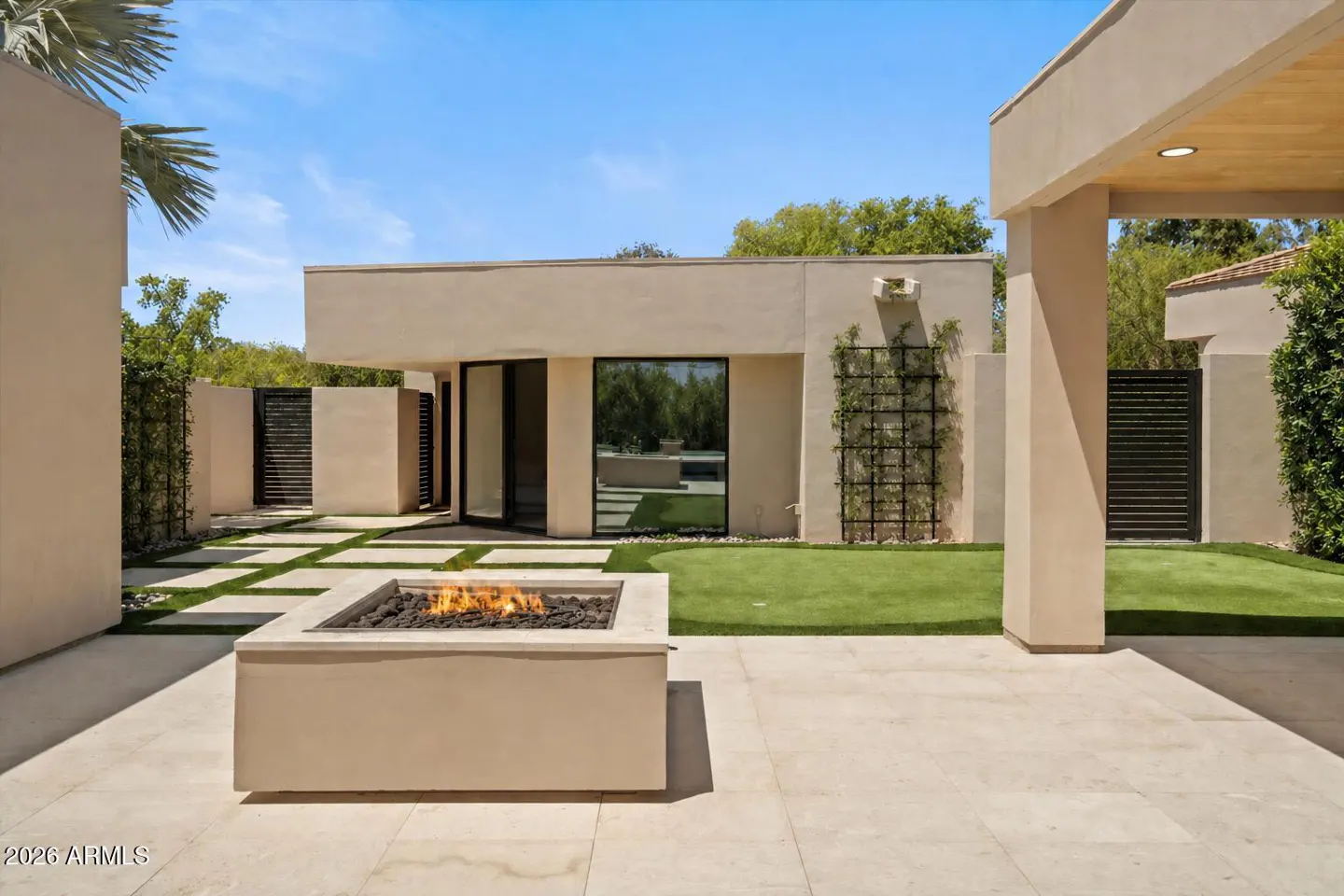 Outdoor patio with a beige fire pit, stone pavers, and green grass. A modern beige building is in the background.