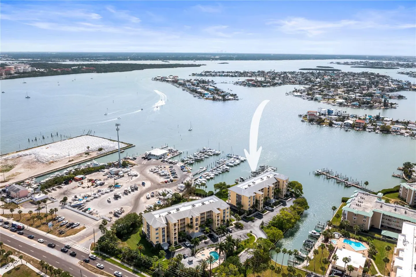 Aerial view of tan condos with a marina, pool, and waterfront access, under a blue sky with an arrow pointing to the condos.