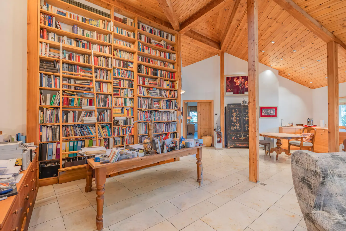 A bright room with a tall wooden bookcase filled with books, a wooden table, and a wooden ceiling.