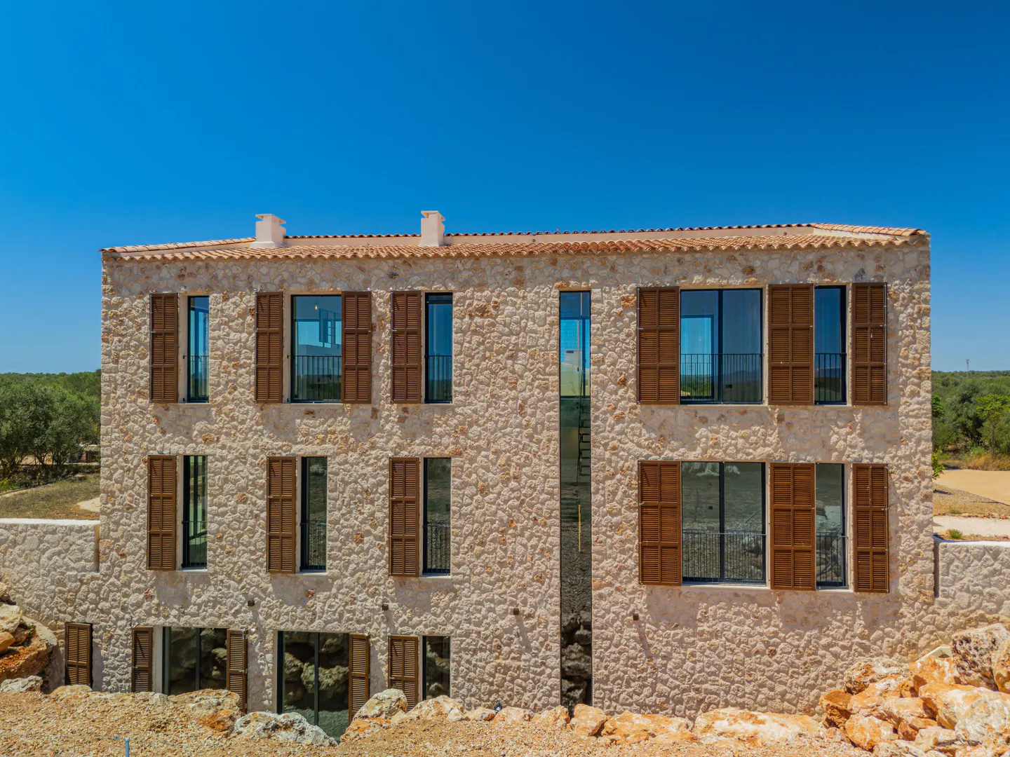 Three-story stone house with brown shutters and a tiled roof under a clear blue sky, surrounded by rocky terrain.