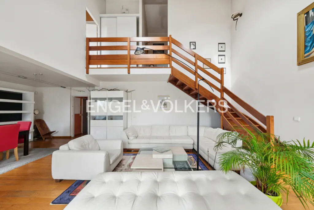 Bright, modern living room with white sofas, wood floors, and a wooden staircase leading to a loft area. A large plant adds a touch of nature.