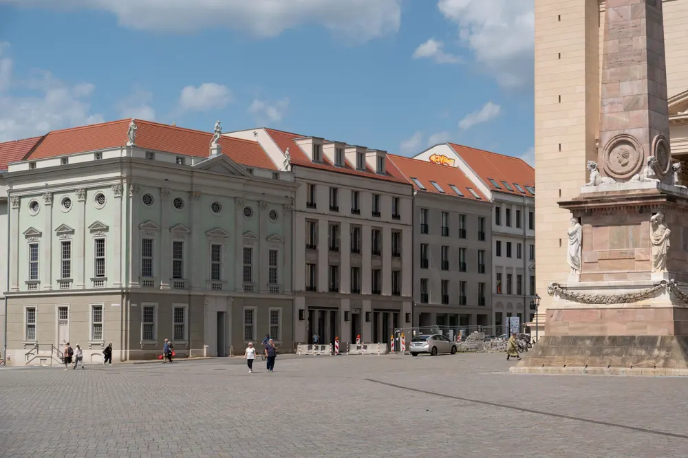 A sunny European square with a stone obelisk in the foreground, historic buildings, and people walking on cobblestones.