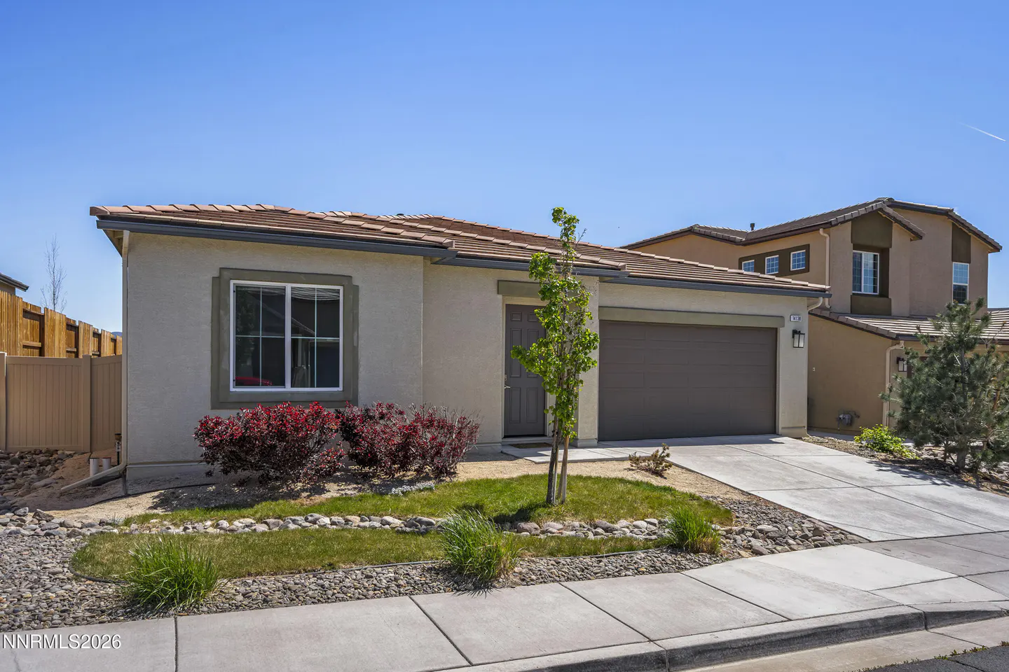 Beige single-story house with a brown roof and garage door under a clear blue sky. Red bushes and green grass accent the front.