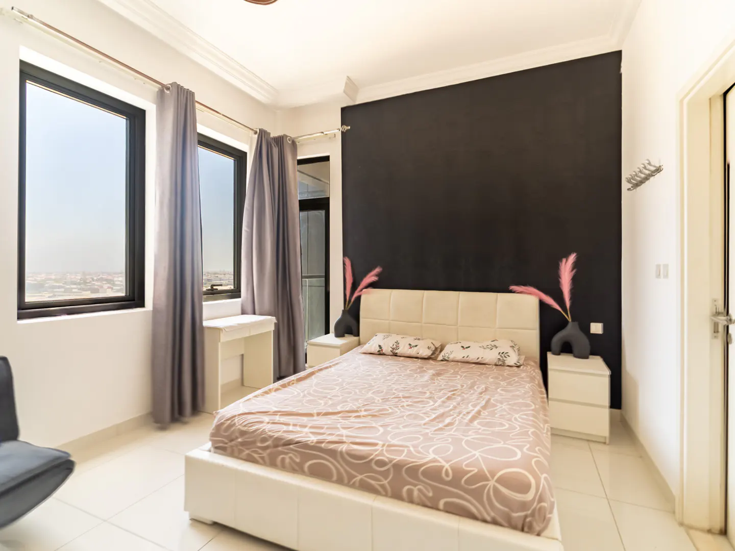 Modern bedroom with a white bed, patterned bedding, black accent wall, grey curtains, and windows with a city view.