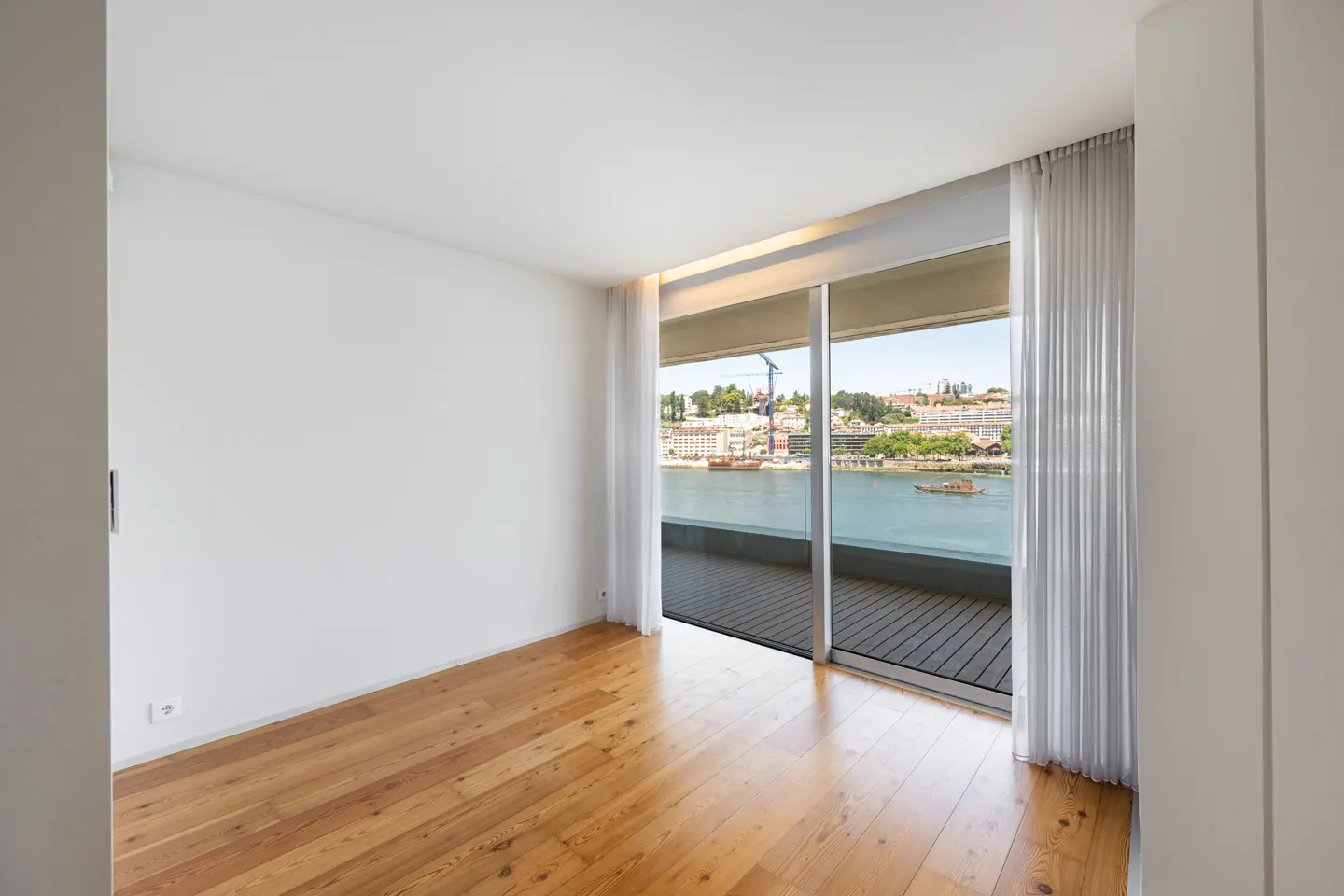 Bright, empty room with wood floors and white walls. Sliding glass doors open to a balcony overlooking a river and city. White curtains frame the doors.