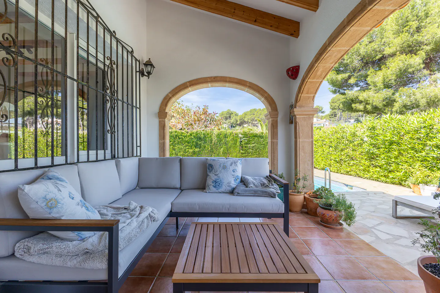 Covered patio with gray sectional sofa, wood table, and arched openings to a green yard with a pool.