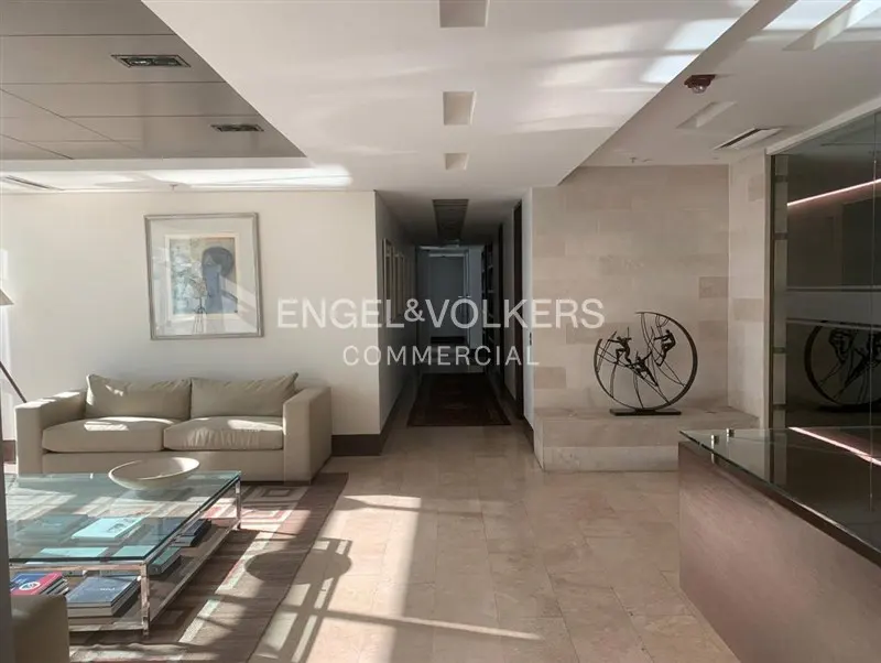 A bright, modern office lobby with a beige sofa, glass table, and a long hallway. The Engel & Volkers logo is visible in the center.