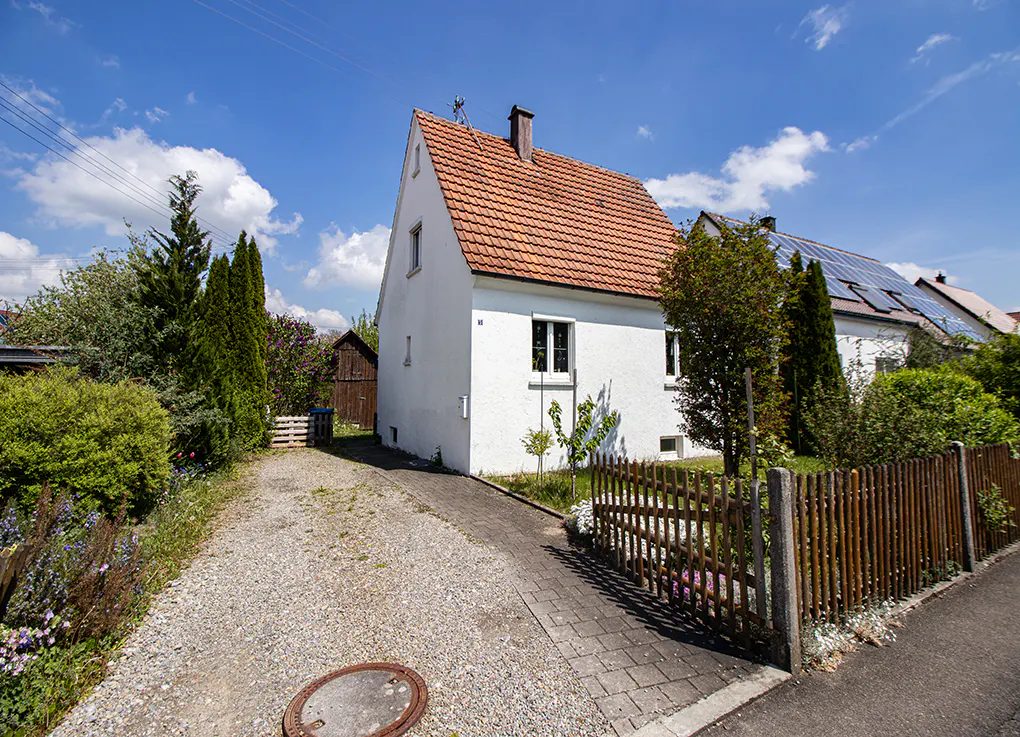 A white house with a red-tiled roof, a gravel driveway, and a wooden fence under a bright blue sky with scattered clouds.