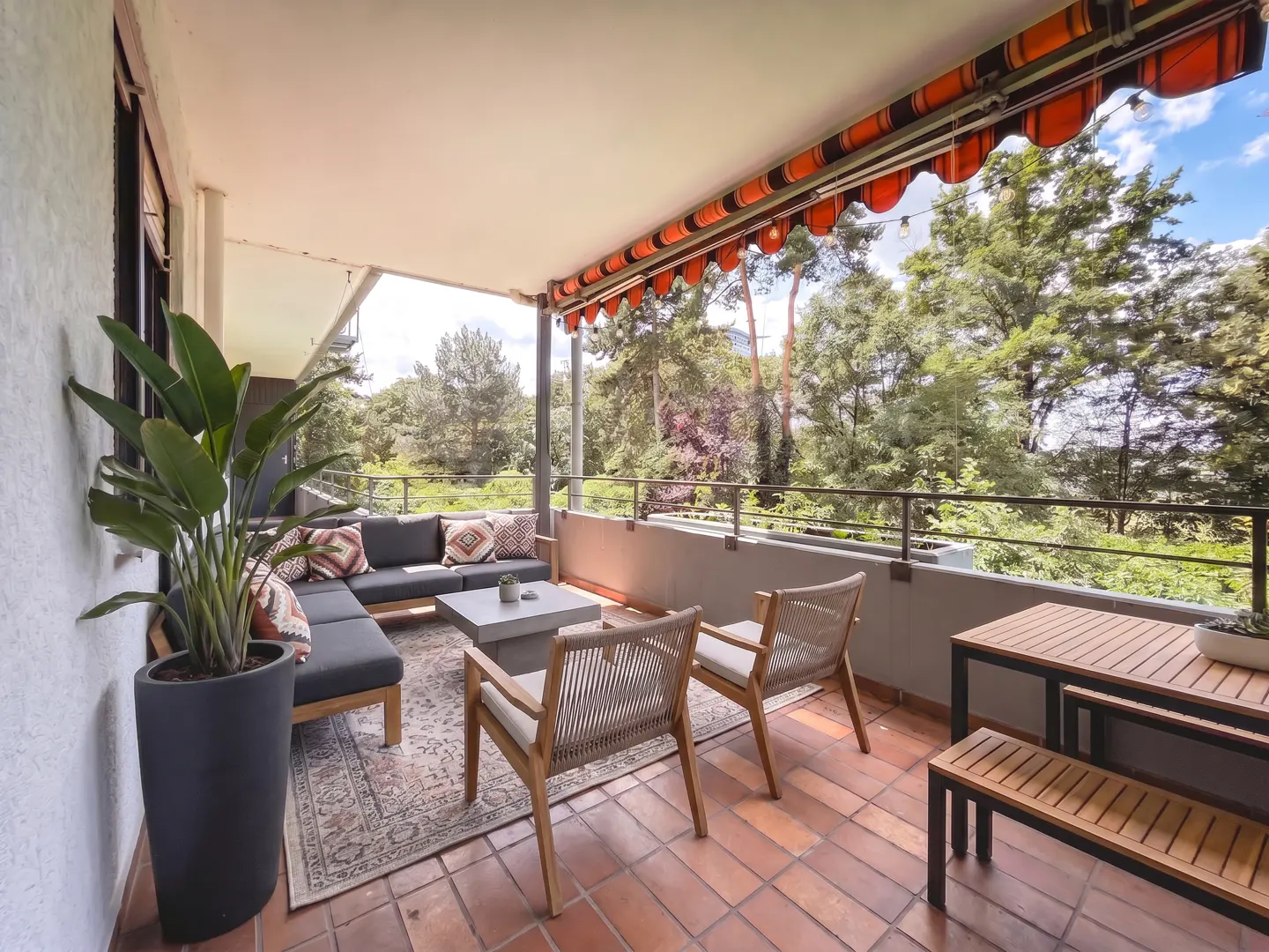 Spacious balcony with terracotta tiles, outdoor sofa, two chairs, dining set, potted plant, and orange striped awning.