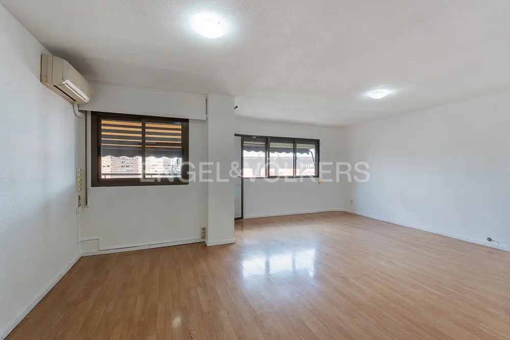 Bright, empty room with light wood floors, white walls, and two windows with brown frames and blinds. An air conditioner is mounted high on the wall.