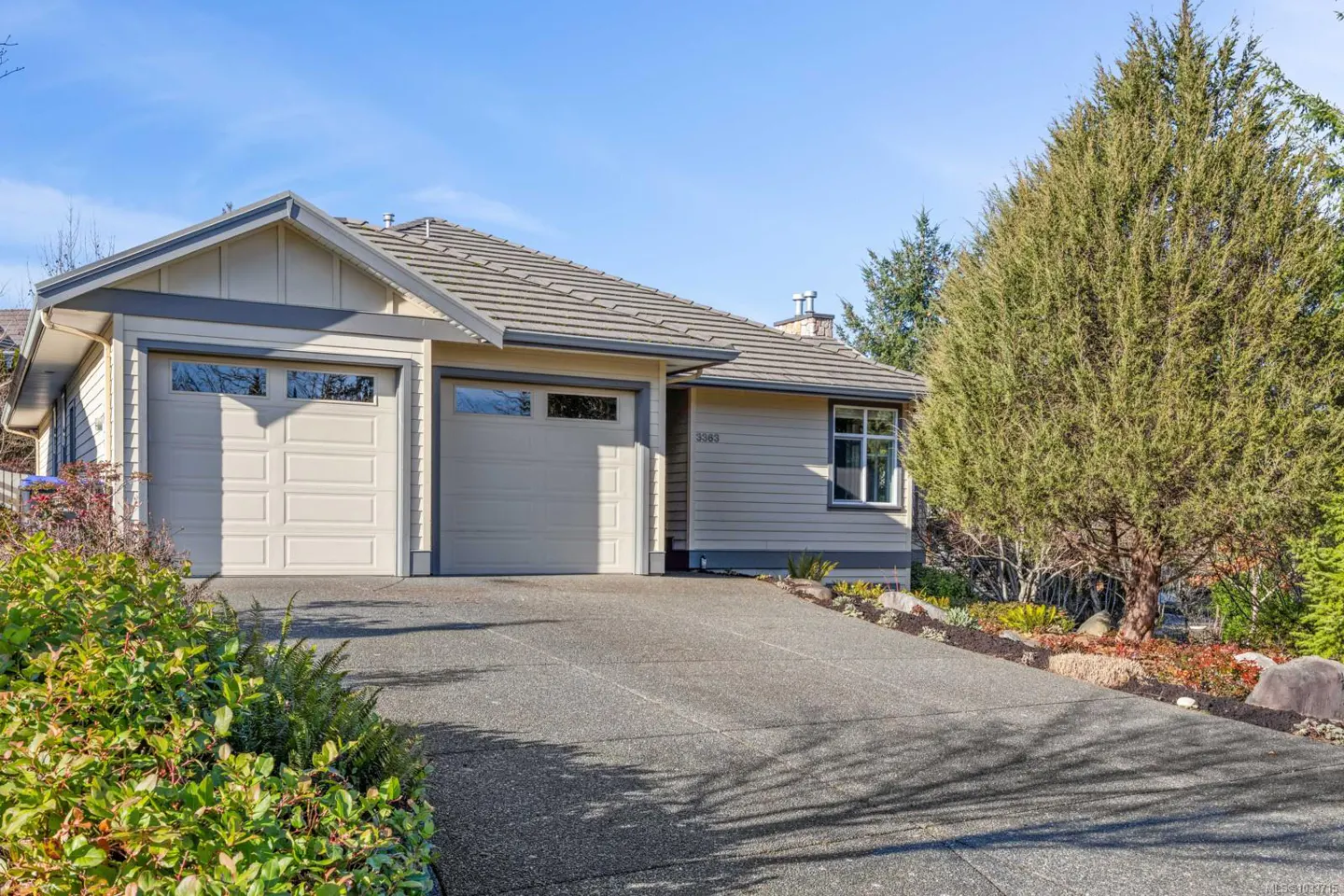 Beige single-story house with a gray roof, two-car garage, and a concrete driveway on a sunny day.