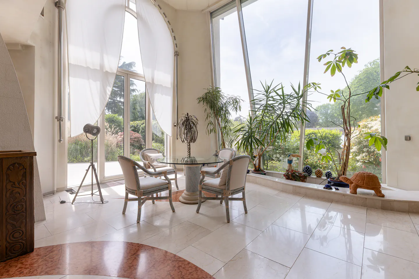 Sunlit dining area with a glass table, four chairs, potted plants, and large windows overlooking a garden.