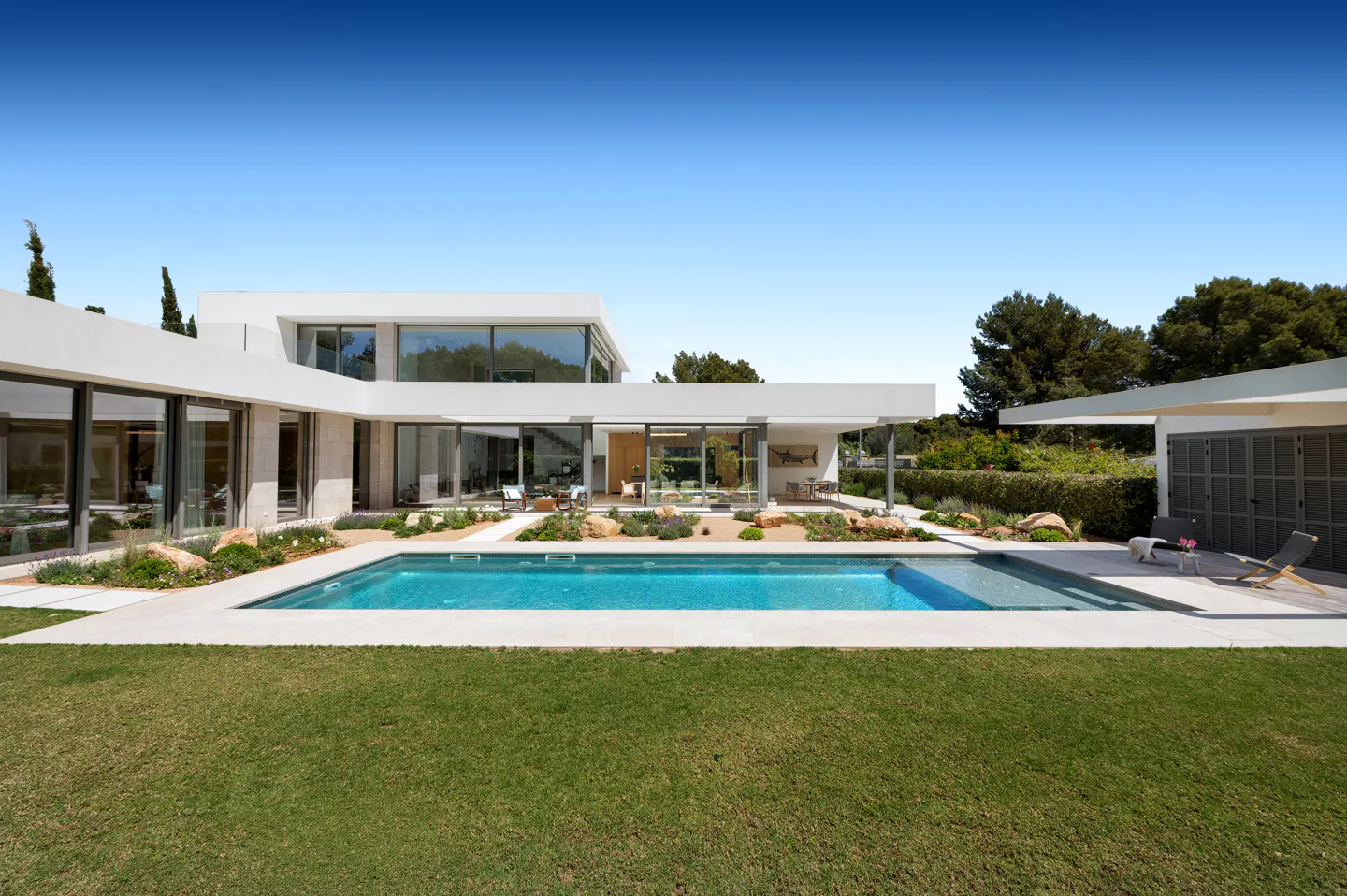 Modern white house with a blue swimming pool, green lawn, and trees under a clear blue sky.