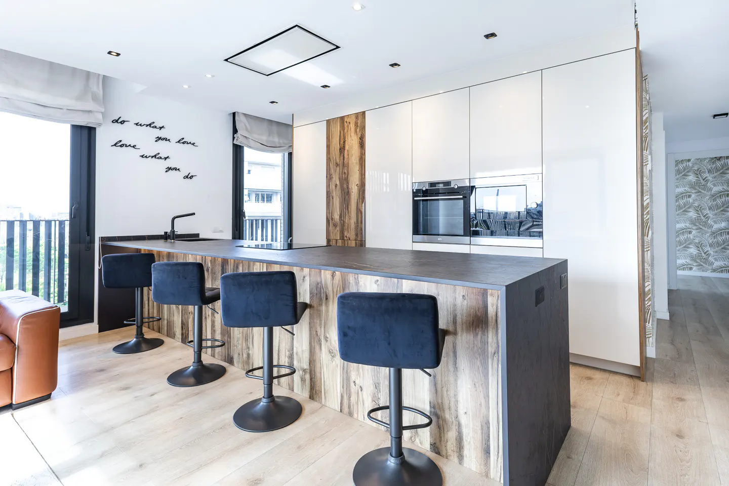 Bright kitchen with wood accents, featuring a dark countertop island with four black stools. White cabinets and a balcony are visible.