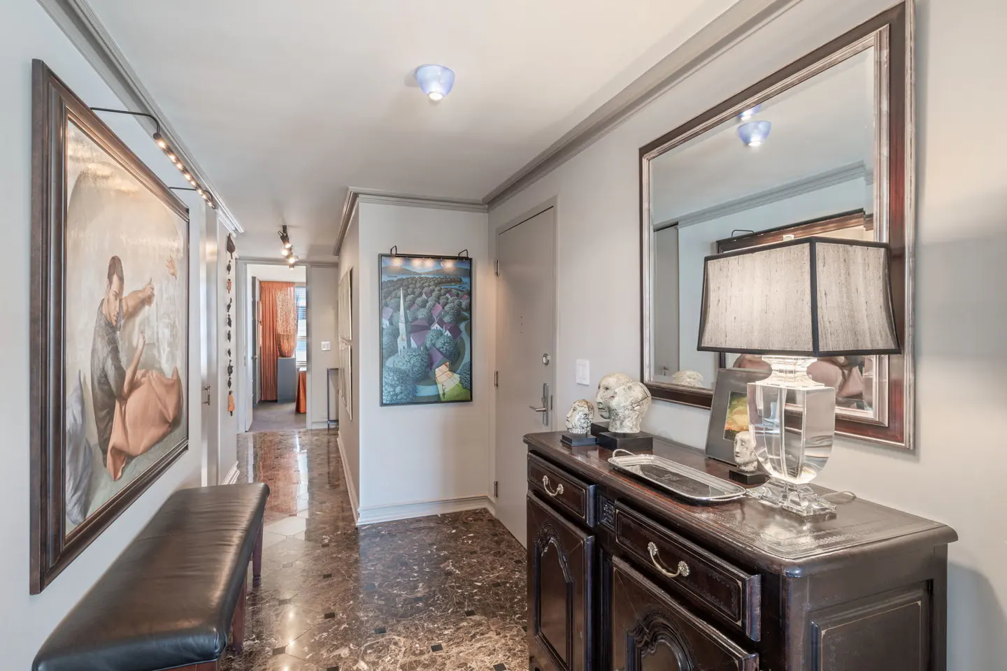 Hallway with marble floors, gray walls, and artwork. A dark wood console table sits beneath a mirror, adorned with a lamp and sculptures.