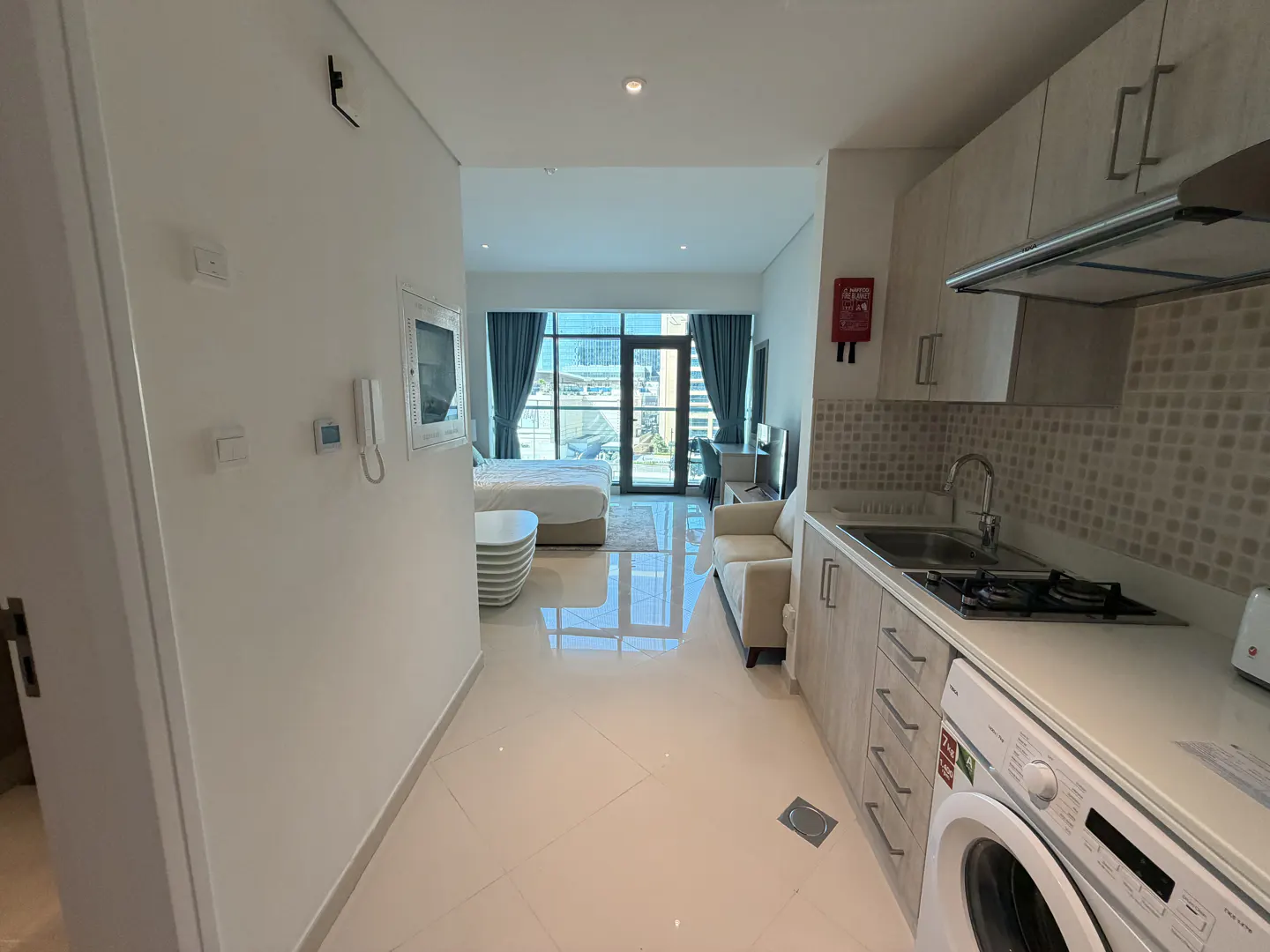 Modern studio apartment with a kitchenette, white tile floors, a sofa, and a bed near a large window with city views.
