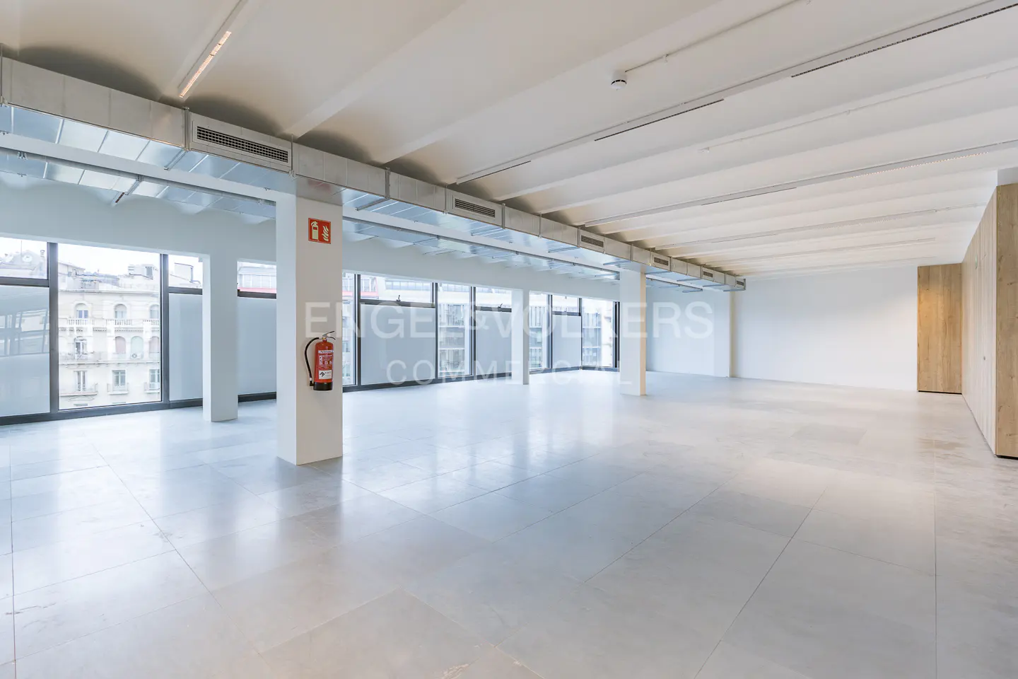 Bright, empty commercial space with white walls, gray floors, and large windows. A red fire extinguisher hangs on a support column.