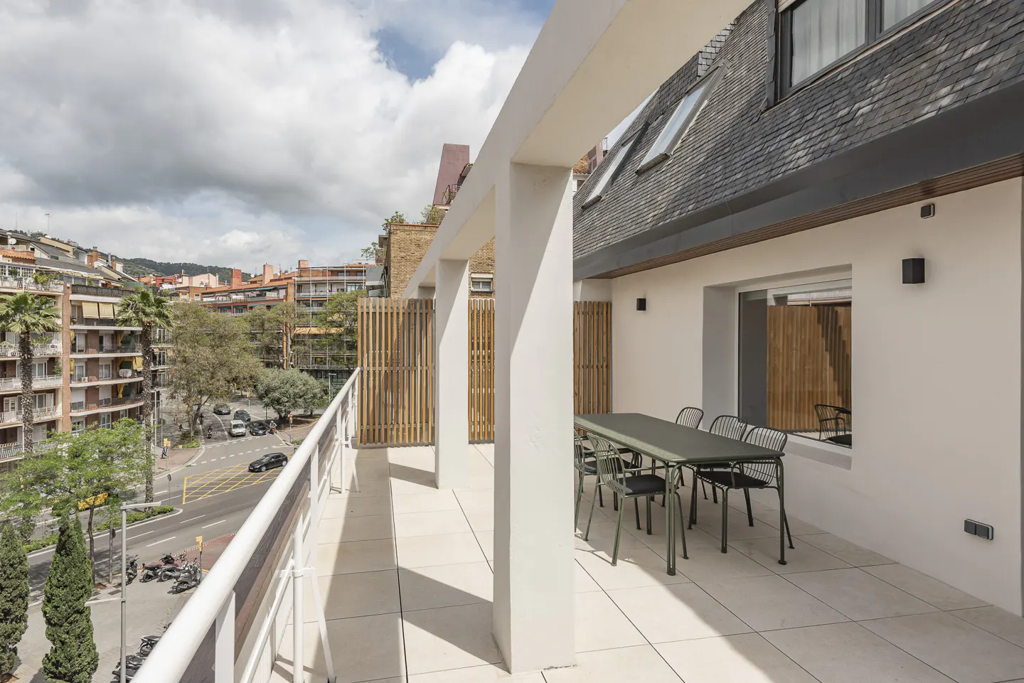 Outdoor patio with a green table and chairs, white walls, and a view of a city street with buildings and trees.
