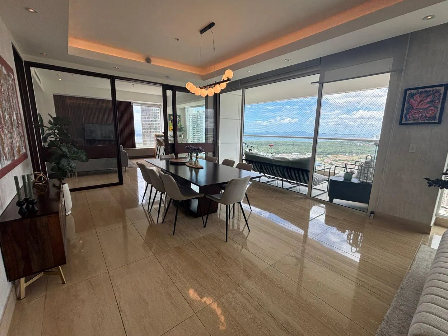 Bright, modern dining room with a dark wood table, light chairs, and a balcony overlooking a scenic view of the ocean and mountains.