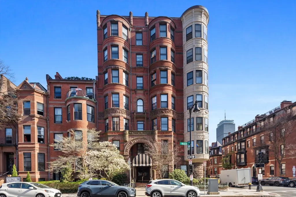 Exterior view of a red brick apartment building with bay windows and a black and white striped awning. Cars are parked on the street.