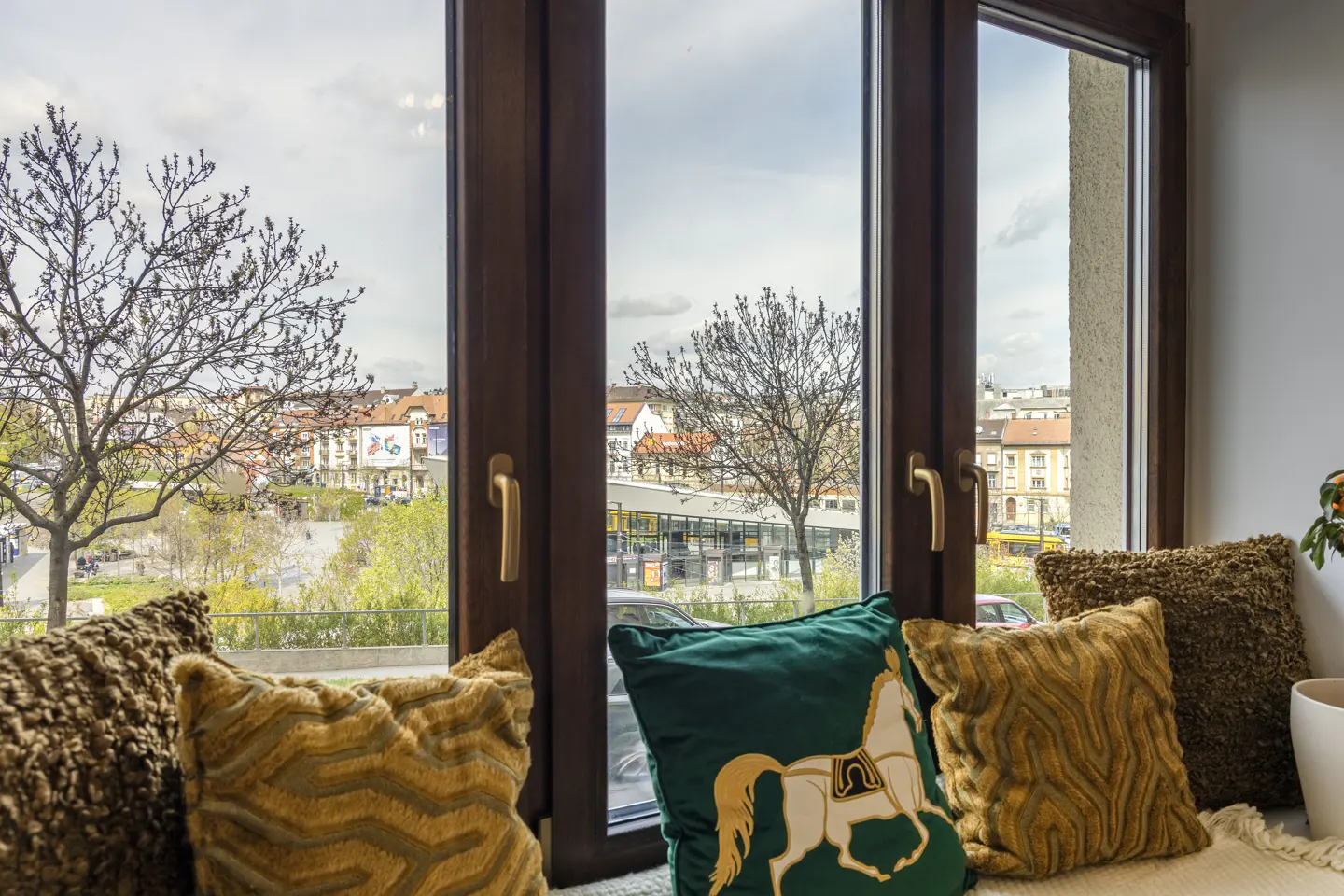 Window seat with brown wood frame, gold handles, and decorative pillows. Outside view of city buildings and trees.