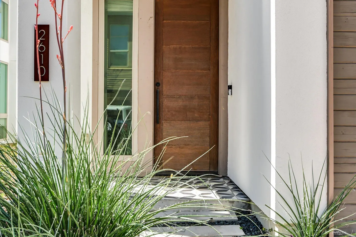 Modern home entrance with a brown wood door, address number 2610, and green plants. The walkway has geometric tiles.