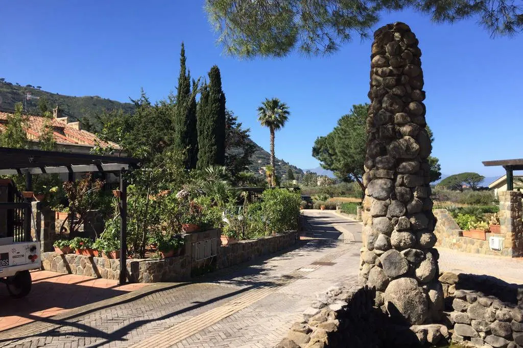 A stone pillar stands beside a brick road lined with lush greenery and trees under a clear blue sky.