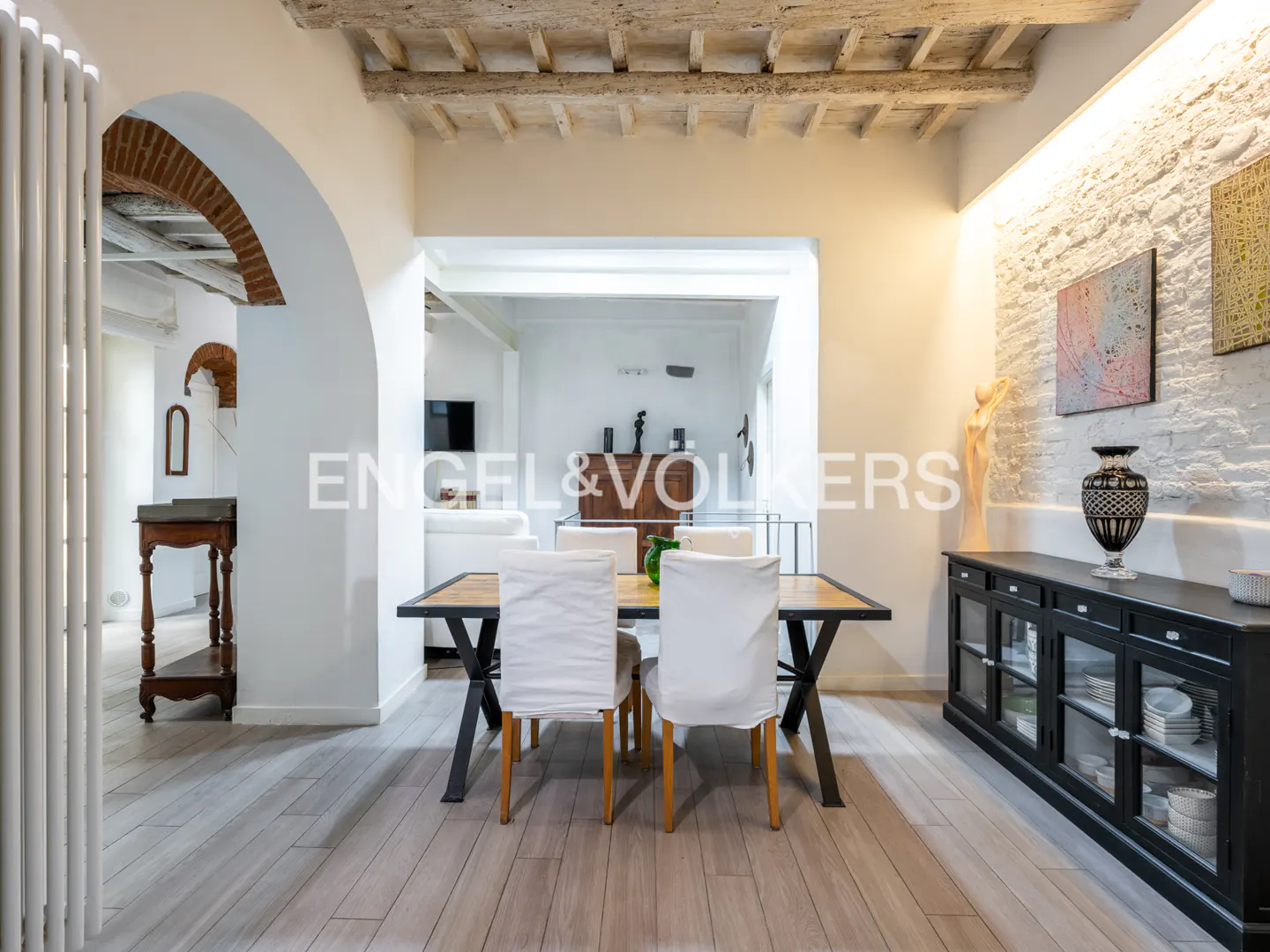 Interior view of a dining room with a wooden table, white chairs, and a black cabinet against a white brick wall.