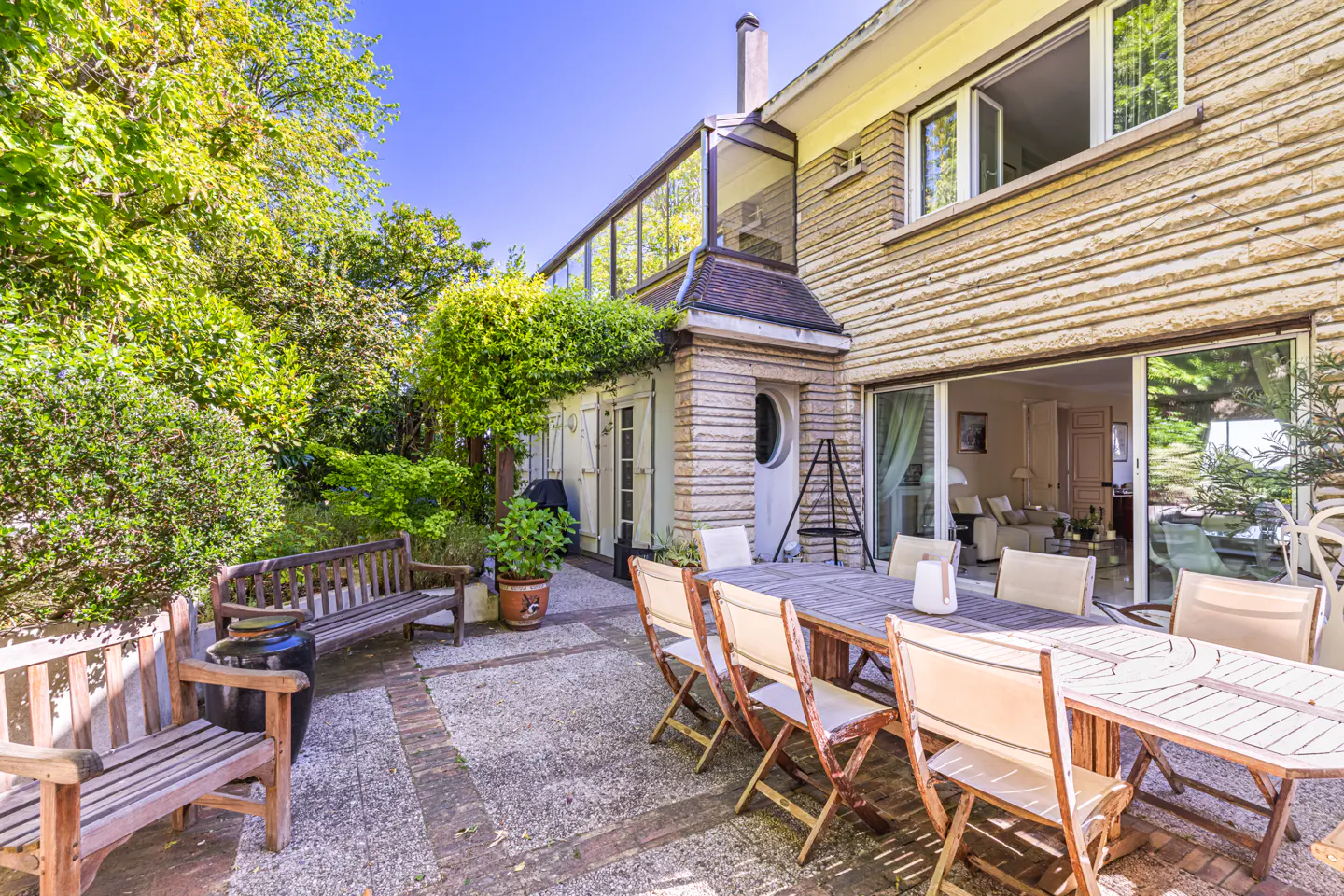 Outdoor patio with a wooden table and chairs, benches, and lush greenery against a stone house with a glass-enclosed upper level.