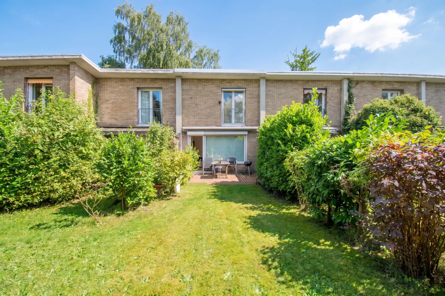 A two-story tan brick townhouse with a lush green backyard, patio furniture, and tall hedges under a bright blue sky.