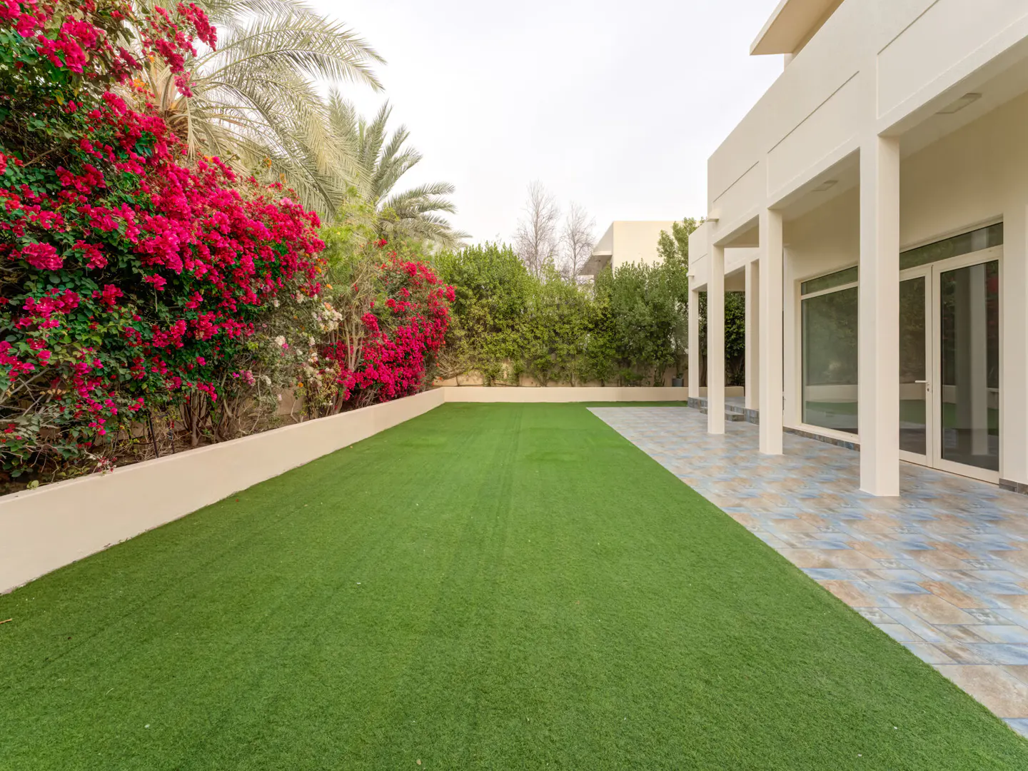 A backyard with green grass, pink flowers, and a covered patio with white pillars.