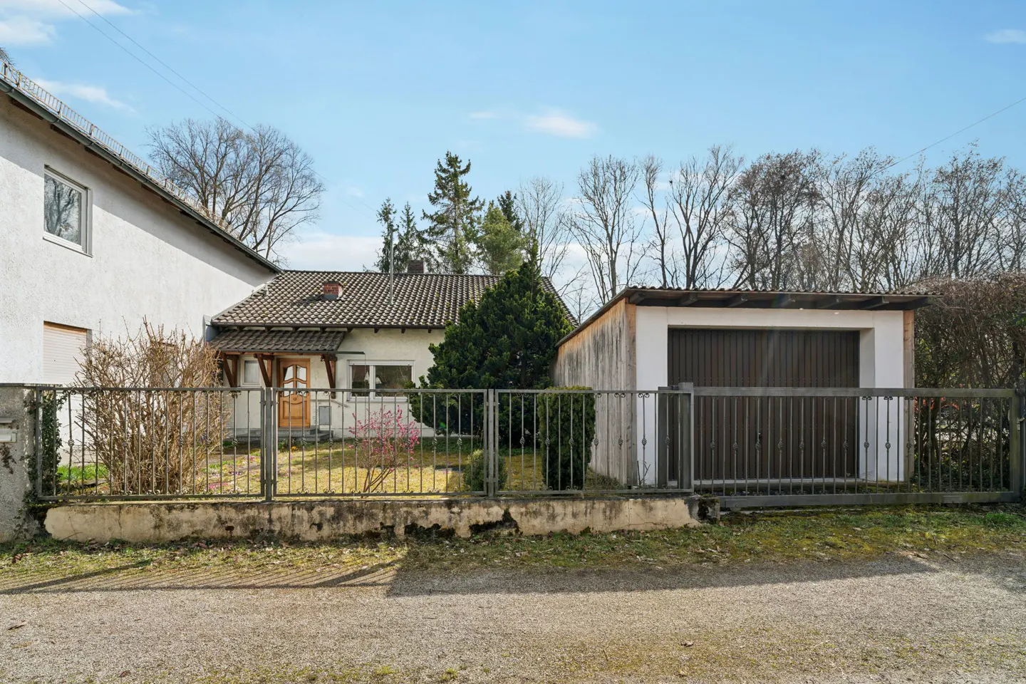 A single-story house with a garage, surrounded by a metal fence and greenery. The house has a brown roof and a wooden door.