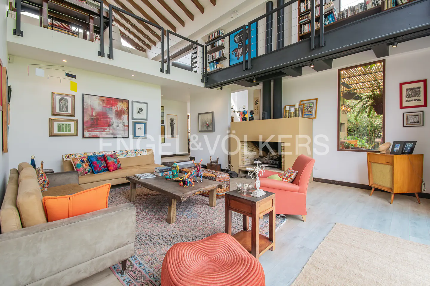 A living room with sofas, a fireplace, and a loft with bookshelves. The room is decorated with art and has a rustic coffee table.