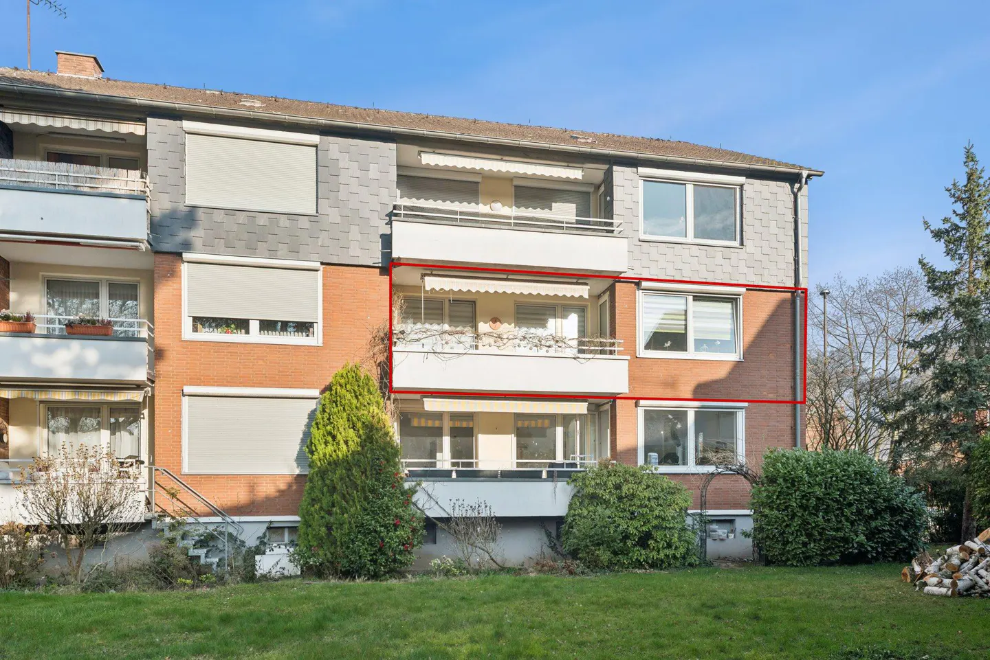 Exterior view of a brick apartment building with white balconies and a red outline. Green lawn and bushes in front. Blue sky.
