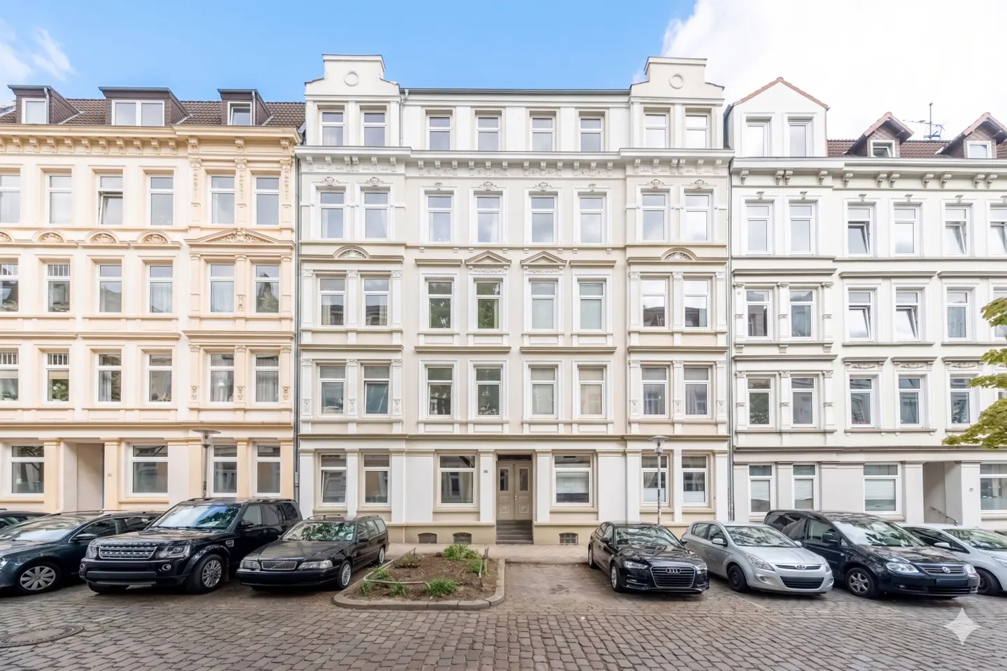 Three adjacent apartment buildings, each with multiple stories and windows, line a cobblestone street with parked cars.