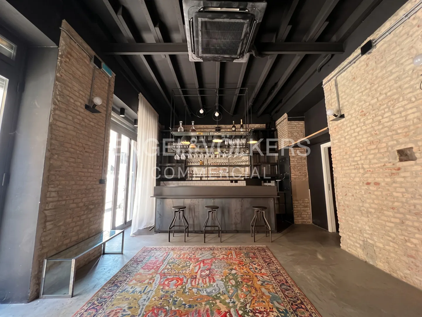 Interior view of a commercial space with a bar, stools, and a colorful rug. Brick walls and a black ceiling add character.