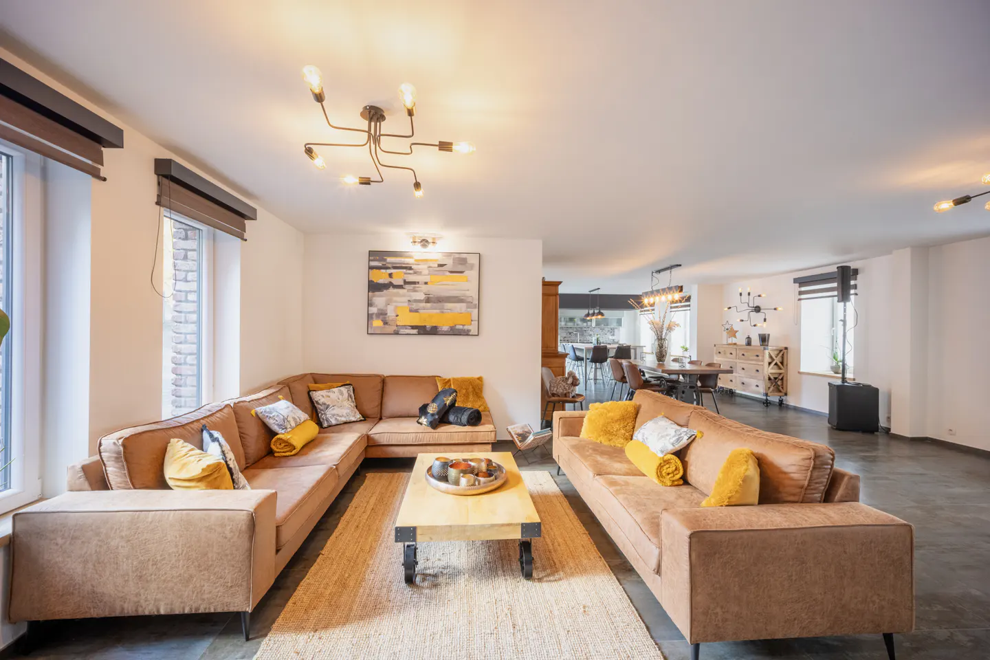 Bright living room with brown sofas, yellow pillows, and a jute rug. A modern light fixture hangs from the white ceiling. Dining area visible in the background.