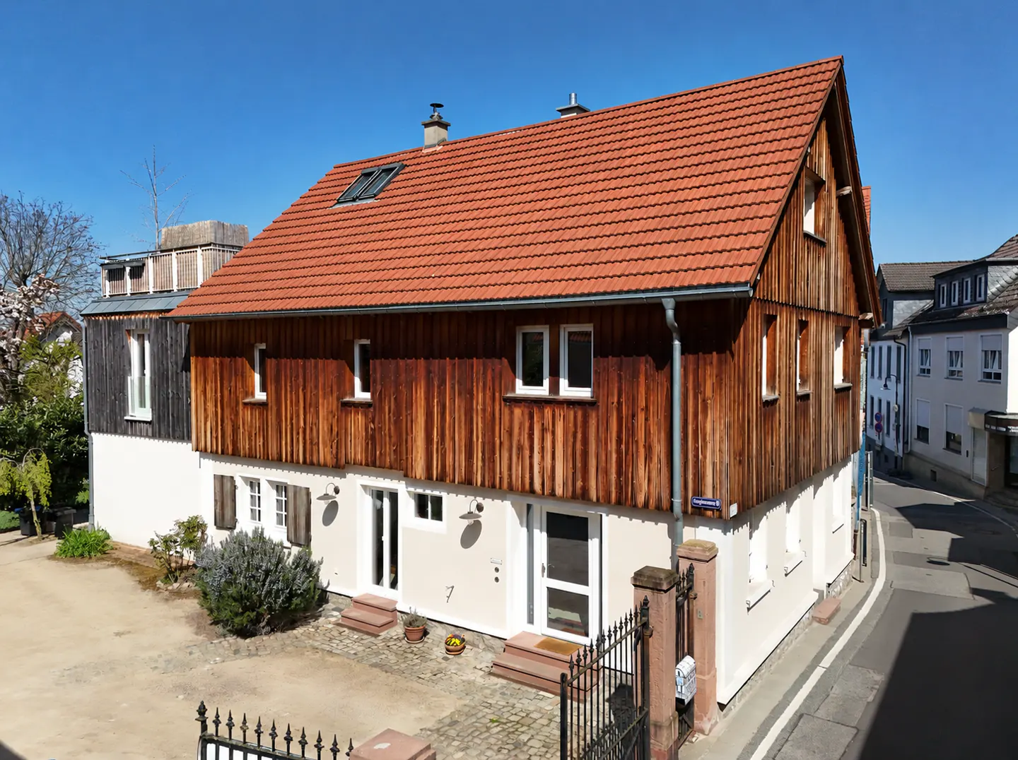 Two-story house with a red tile roof, wood siding, and a white stucco base. A black iron fence is in the foreground.