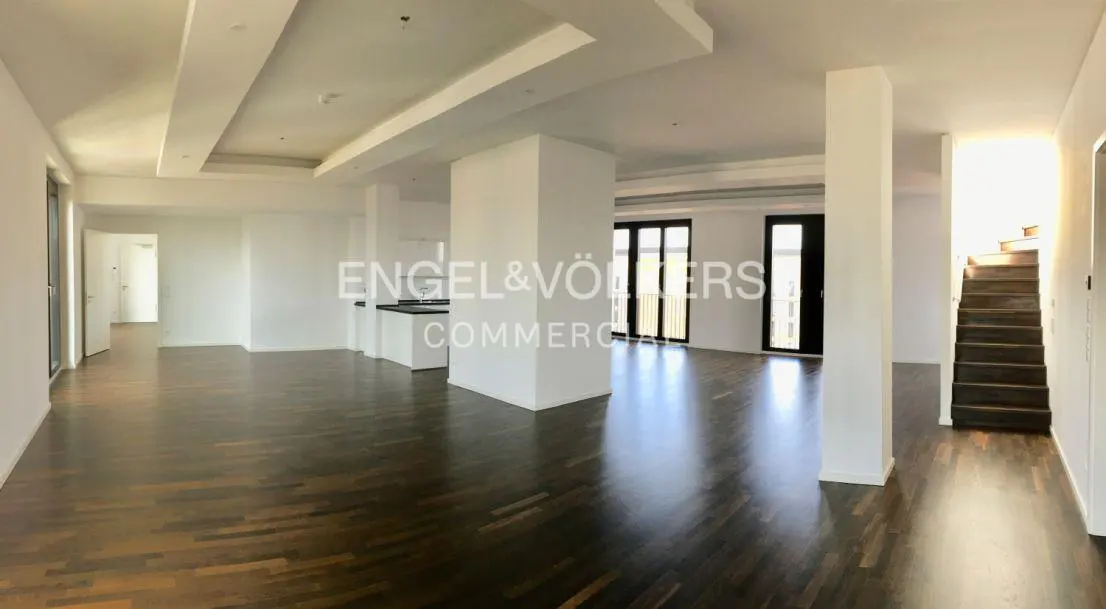 A bright, open-concept space with dark wood floors, white walls, and support columns. A staircase is visible on the right.