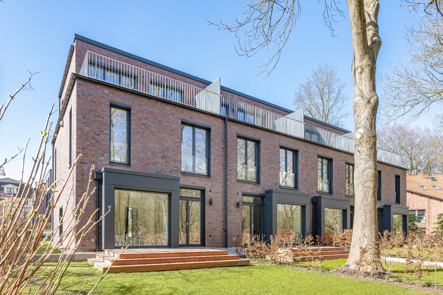 Exterior view of a three-story brick townhouse with black trim, large windows, and a rooftop balcony. A green lawn and trees surround the building.