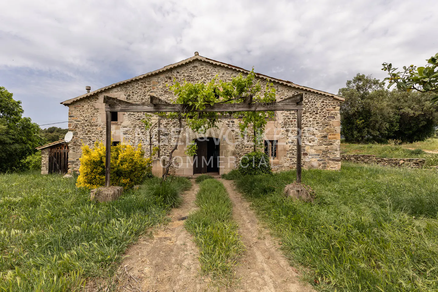 Stone house with a wooden pergola covered in vines, leading to a dark doorway, surrounded by green grass and trees.