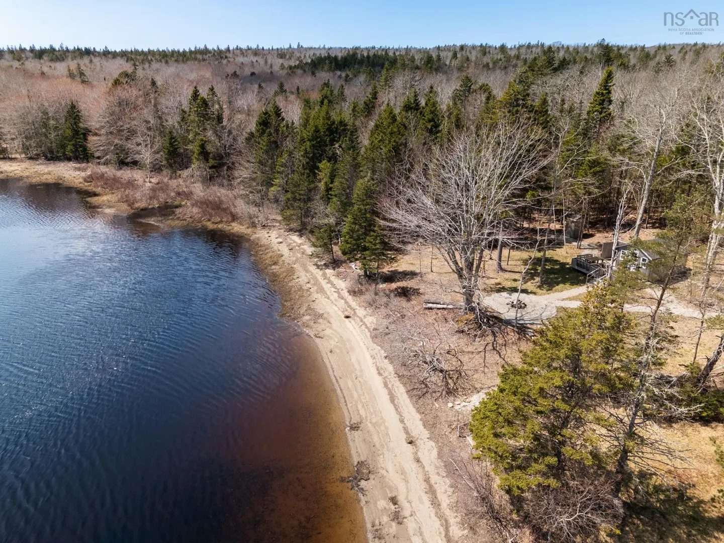 Aerial view of a sandy beach bordering a dark blue lake and a forest with a small house visible in the trees.