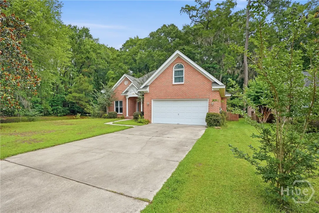 Single-story red brick house with a white garage door, concrete driveway, and lush green trees in the background.