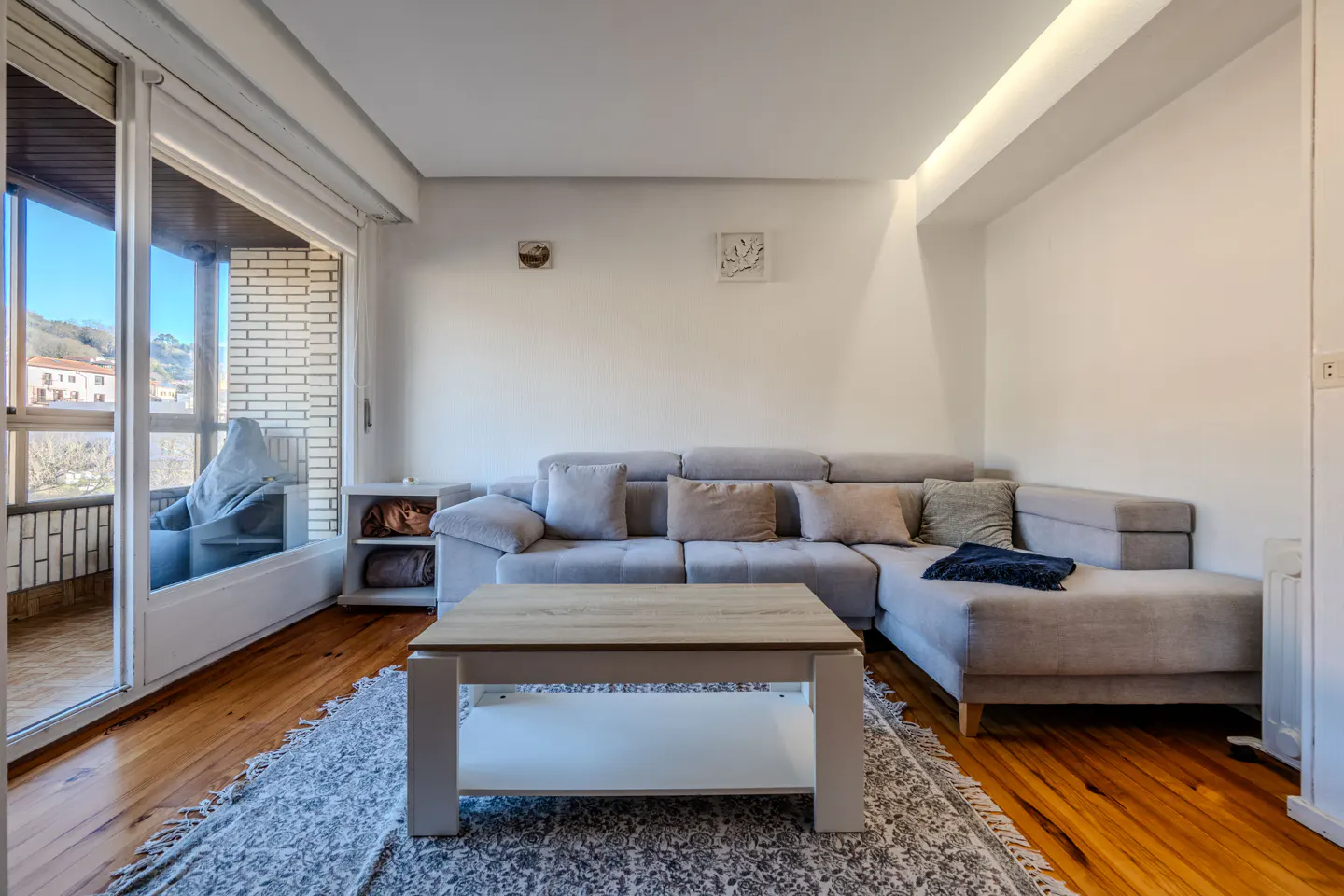 Living room with a gray sectional sofa, pillows, and a wooden coffee table on a patterned rug. Balcony with a view is visible.