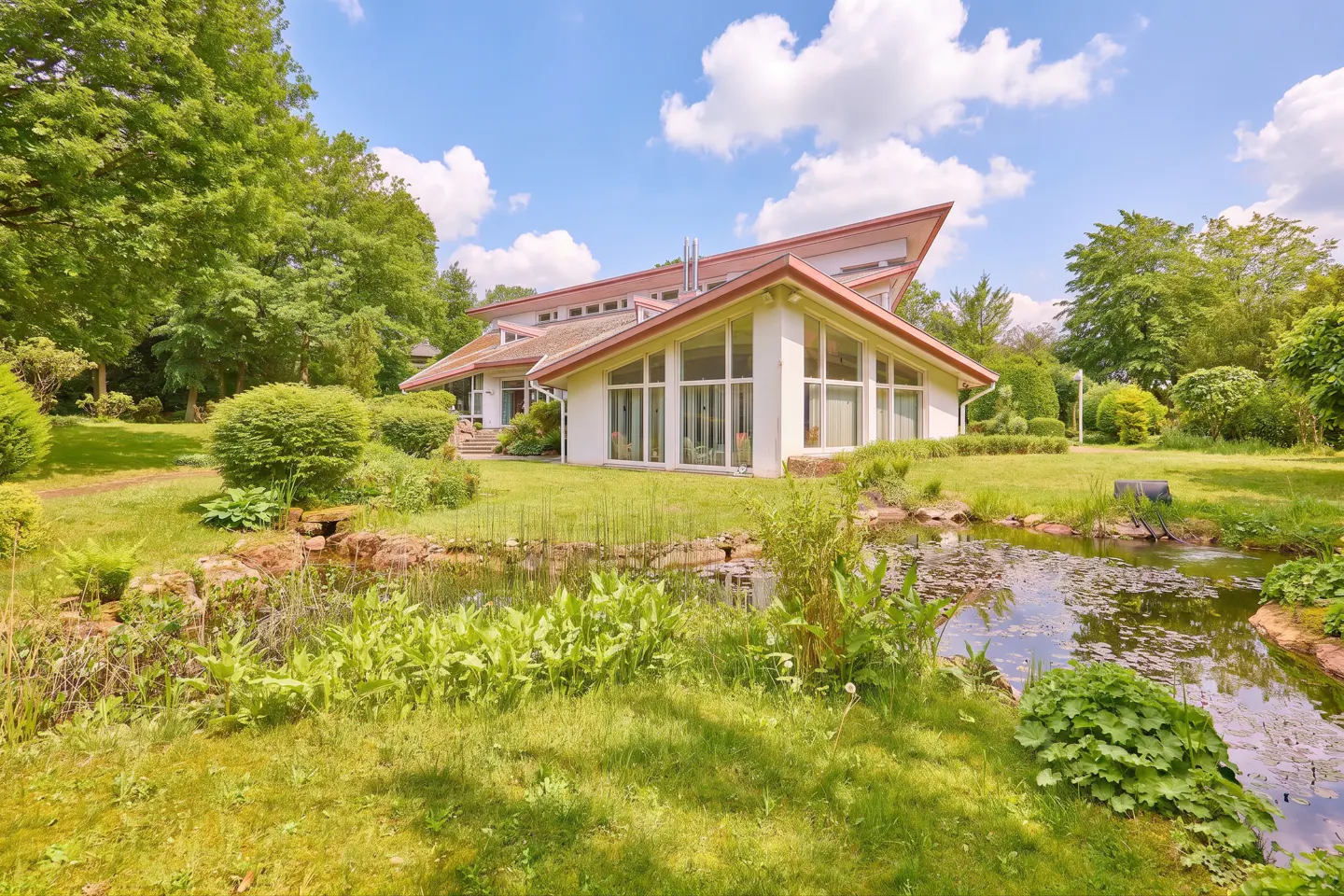 Exterior view of a modern white house with a red roof, surrounded by green trees and a pond with lily pads.