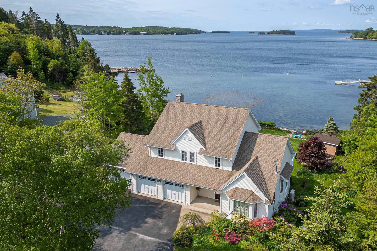 Aerial view of a white house with a brown roof, surrounded by green trees and blue water.