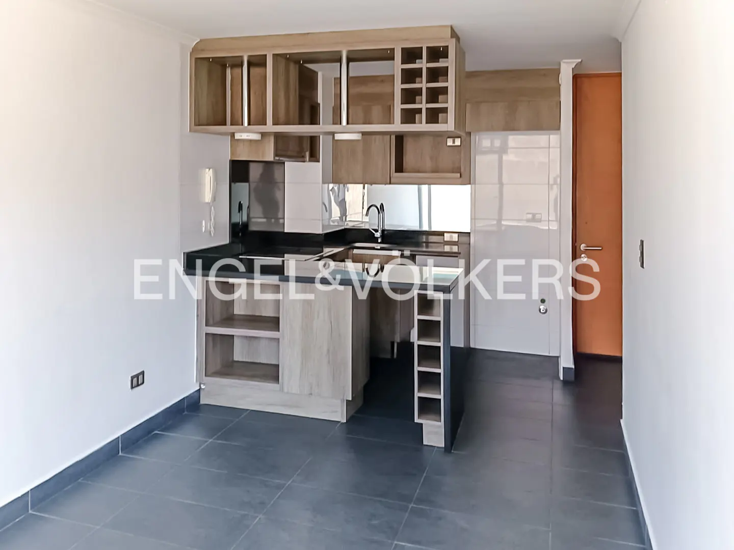 A modern kitchen with light wood cabinets, black countertops, and gray tile flooring. The Engel & Volkers logo is superimposed on the image.