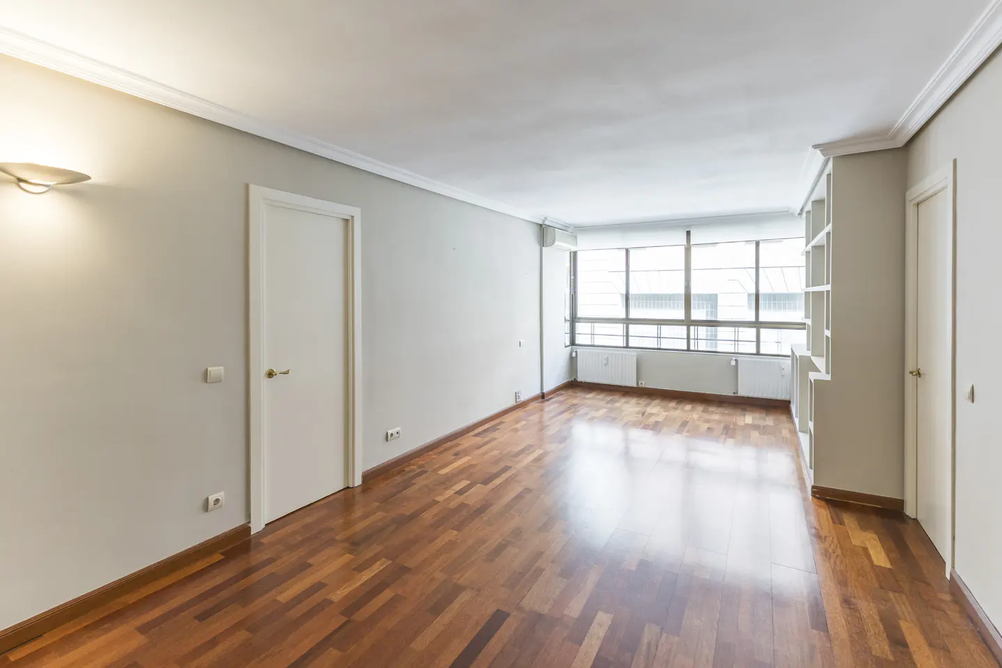 Empty room with hardwood floors, white walls, and a large window. There are two white doors and a built-in bookshelf.
