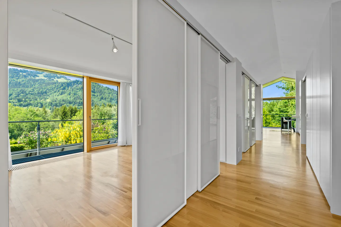 Modern hallway with light wood floors, white sliding doors, and large windows overlooking a lush green mountain landscape.