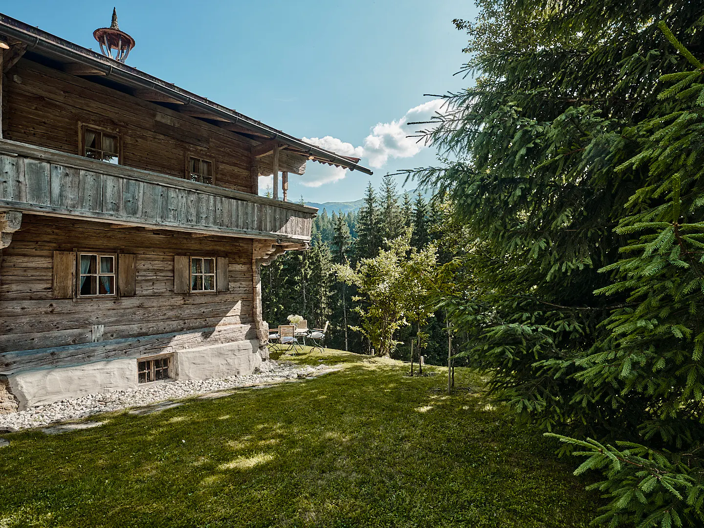 Exterior view of a rustic wooden chalet with a balcony, surrounded by green lawn and trees on a sunny day.